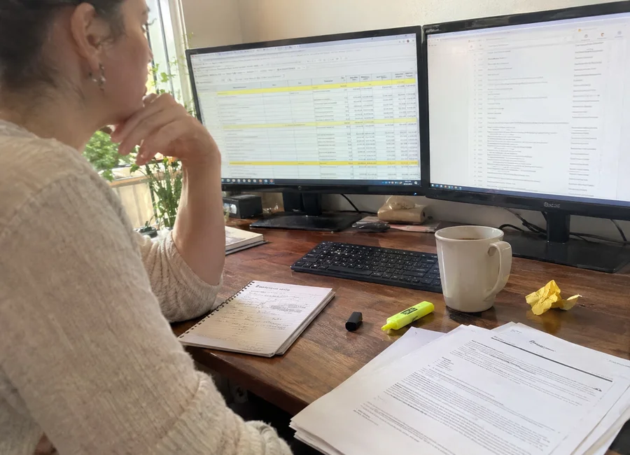 Home office desk with monitors showing a property criteria spreadsheet, printed lease pages, and a notepad