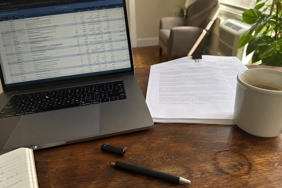 Laptop open to a property criteria spreadsheet beside a printed lease and a coffee cup